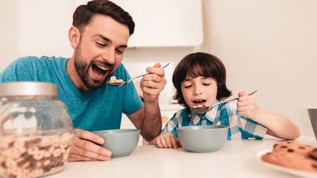 Smiling Son and Father Have Breakfast in Kitchen. Boy in Shirt. Modern Kitchen. Sitting Boy. Boy with Spoon. Breakfast in Morning. White Table in Kitchen. Gray Bowl on Table. Young Father.の写真素材