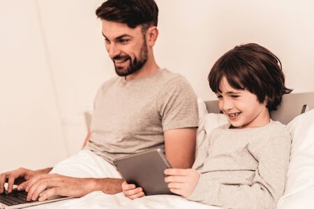Son with Father Using Laptop in Bed in Morning. Digital Device. Happy Family Concept. White Bed in Room.の写真素材