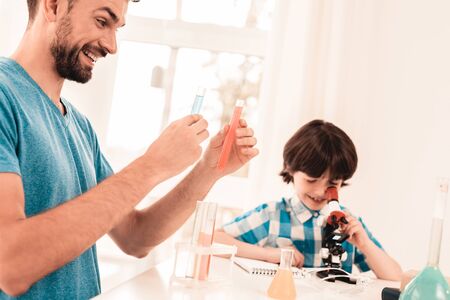 Youn Bearded Father Teaching Son in Shirt at Home. Education at Home. Using Microscope. Studding Chemistry. White Table in Room. Sitting Boy. Young Father. Lesson at Home. Education Concept.の写真素材