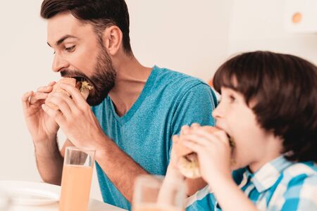 Smiling Son and Father Have Lunch in Kitchen. Boy in Shirt. Modern Kitchen. Sitting Boy. Boy with Spoon. Breakfast in Morning. White Table in Kitchen. Burger in Hands. Young Father.の写真素材