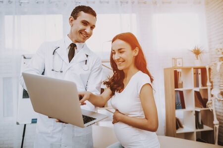 Pregnant Girl at the Gynecologist Doctor. Doctor is a Man. Doctor is Showing Something on Laptop to Woman. Girl is Sitting on Couch and Pointing on Laptop Screen. Persons is Smiling. People at Clinic.の写真素材