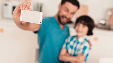 Happy Father and Son Using Smartphone at Home. Smiling Boy. Modern Kitchen. Sitting Boy. Boy in Shirt. Breakfast in Morning. White Table in Kitchen. Using Digital Device. Happy Family.の写真素材