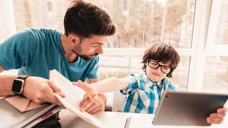 Father Trying to Help Son in Glasses with Homework. Education at Home. White Table in Room. Boy Using Digital Divece. Young Father. Lesson at Home. Education Concept. Boy in Shirt. Playing on Tablet.の写真素材