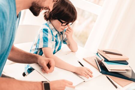 Little Boy in Glasses Doing Homework with Father. Education at Home. White Table in Room. Sitting Boy. Young Father. Lesson at Home. Education Concept. Writing in Notebook. Boy in Shirt.の写真素材