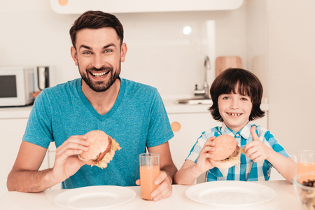 Smiling Son and Father Have Lunch in Kitchen. Boy in Shirt. Modern Kitchen. Sitting Boy. Boy with Spoon. Breakfast in Morning. White Table in Kitchen. Burger in Hands. Young Father.の写真素材