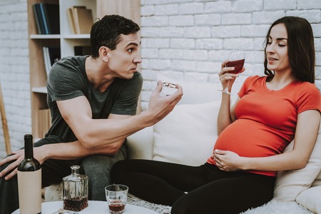 Man is Argues with His Wife. Wife is a Young Pregnant Girl. Girl is Drinking a Wine and Smiling. People is Sitting on Couch. Man is Showing a Cigarettes to Woman. Persons Located at Home.の写真素材