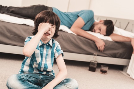 Little Boy Sitting in Room with Drunken Father. Crisis in Family. Alcoholism Problem Concept. Glass of Whiskey. Young Boy in Shirt. Modern Social Problems Concept. Sitting Sad Boy.の写真素材
