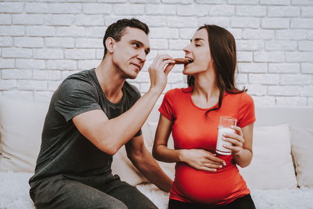 Couple is Sitting on Couch. Man is Feeding a Woman a Cookie. Woman is a Young Pregnant Girl. Girl is Holding a Cup with Milk. Woman is Touching Her Belly and Smiling. People Located at Home.の写真素材