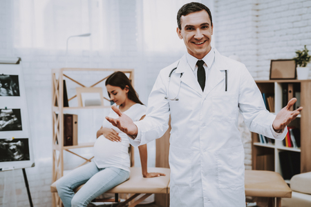 Pregnant Girl at the Gynecologist Doctor. Doctor is a Young Man. Doctor is Smiling into a Camera and Gesticulating. Woman is Sitting on Couch and Touching Her Belly. People Located at Clinic.の写真素材