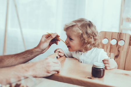 Man Spends Time with His Son. Father of Boy is Engaged in Raising Child. Father is Feeding His Blonde Son from Spoon. People is Sitting on Chairs at Table. People Located on Kitchen.の写真素材