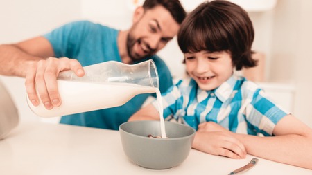 Smiling Son and Father Have Breakfast in Kitchen. Boy in Shirt. Modern Kitchen. Sitting Boy. Boy with Spoon. Breakfast in Morning. White Table in Kitchen. Gray Bowl on Table. Young Father.の写真素材
