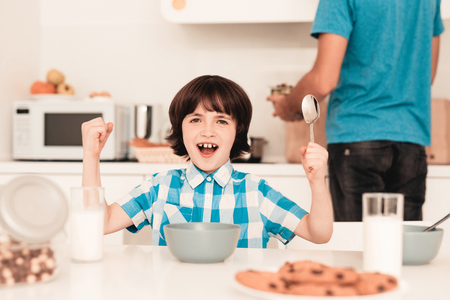 Little Boy in Shirt Have Fun in Kitchen in Morning. Son with Father. Modern Kitchen. Sitting Boy. Boy with Spoon. Breakfast in Morning. White Table in Kitchen. Gray Bowl. Young Father.の写真素材
