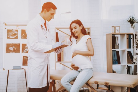 Pregnant Girl at the Gynecologist Doctor. Doctor is Young Man. Doctor is Writing Information About a Patient. Girl is Sitting on Couch and Touching Her Belly. Girl is Smiles. People Located at Clinic.の写真素材