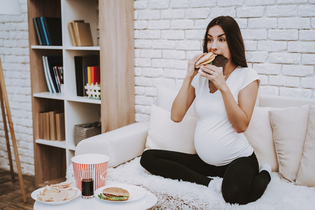Woman is Eating a Hamburger and Chocolate Bar Together. Woman is a Young Brunette Pregnant Girl. Girl is Sitting on Couch. Different Food and Cola on Table. Person is Located at Home.の写真素材