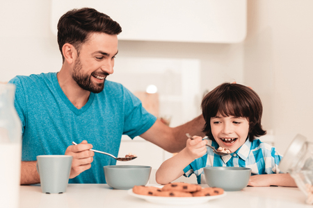 Smiling Son and Father Have Breakfast in Kitchen. Boy in Shirt. Modern Kitchen. Sitting Boy. Boy with Spoon. Breakfast in Morning. White Table in Kitchen. Gray Bowl on Table. Young Father.の写真素材