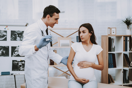 Pregnant Girl at the Gynecologist Doctor. Doctor is a Young Smiling Man. Doctor is Showing the Pregnancy Test to Woman. Girl is Sitting on Couch and Touching Her Belly. People Located at Clinic.の写真素材
