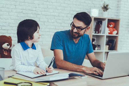 Man Spends Time with His Son. Father of Boy is Engaged in Raising Child. Father is Doing a Homework with Son. Man is Typing in Laptop. People is Sitting at Table. People Located at Home.の写真素材