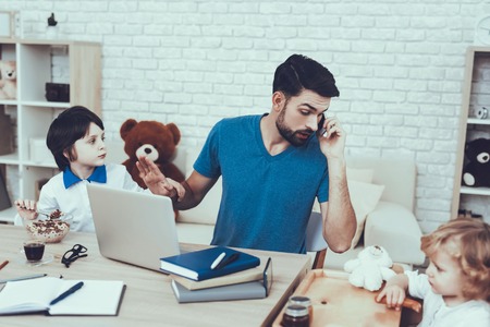 Man Spends Time with His Sons. Father is Engaged in Raising Children. Eldest Son is Eating a Breakfast. Father is Working and Talking on Cellphone. People is Sitting at Table. People Located at Home.の写真素材