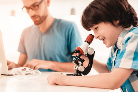 Youn Bearded Father Teaching Son in Shirt at Home. Education at Home. Using Microscope. Studding Chemistry. White Table in Room. Sitting Boy. Young Father. Lesson at Home. Education Concept.の写真素材