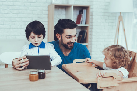 Man Spends Time with His Sons. Father of Boys is Engaged in Raising Children. Eldest Son is Watching Video on Tablet PC. Father is Smiling. Boys is Sitting at Table. People Located on Kitchen.の写真素材