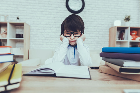 Child is Doing Homework. Child is a Little Black Haired Boy. Boy Wearing a Glasses. Boy is Upset. Child is Sitting at Table. Books and Mobile Phone on Table. Boy is Located at Home.の写真素材