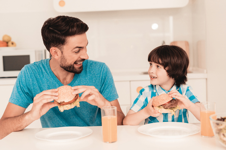 Smiling Son and Father Have Lunch in Kitchen. Boy in Shirt. Modern Kitchen. Sitting Boy. Boy with Spoon. Breakfast in Morning. White Table in Kitchen. Burger in Hands. Young Father.の写真素材
