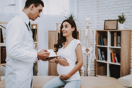 Pregnant Girl at the Gynecologist Doctor. Doctor is Young Man. Doctor is Writing Information About a Patient. Girl is Sitting on Couch and Touching Her Belly. Girl is Smiles. People Located at Clinic.の写真素材