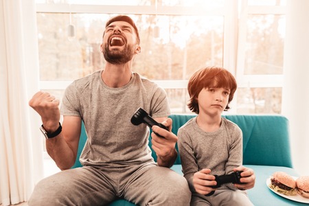 Father and Son Sitting and Playing on Console. Sitting Boy. Young Father. Playing on Console. Joystick in Hands. Young Bearded Man at Home. Sitting on Blue Sofa. Happy Family Concept.の写真素材