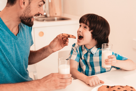 Smiling Son and Father Have Breakfast in Kitchen. Boy in Shirt. Modern Kitchen. Sitting Boy. Boy with Spoon. Breakfast in Morning. White Table in Kitchen. Gray Bowl on Table. Young Father.の写真素材