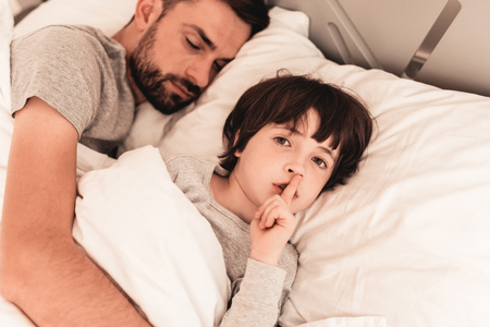 Little Son with Tired Sleeping Father in Bed. Family in Morning. Happy Family Concept. White Bed in Room. Father and Son at Home. Morning Routine. Lying in Bed. Sleeping Man. Blanket and Pillows.の写真素材