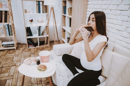 Woman is Eating a Hamburger and Chocolate Bar Together. Woman is a Young Brunette Pregnant Girl. Girl is Sitting on Couch. Different Food and Cola on Table. Person is Located at Home.の写真素材