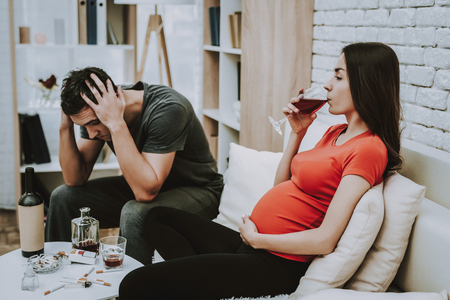 Man is Shocked by His Wife. Wife is a Young Pregnant Girl. Girl is Drinking a Wine. Woman is Touching Her Belly. People is Sitting on Couch. Cigarettes and Alcohol on Table. Persons Located at Home.の写真素材