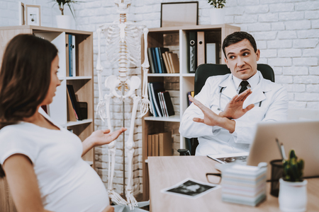 Pregnant Girl at the Gynecologist Doctor. Doctor is a Young Man. Woman is Touching Her Belly and Gesticulating. Doctor is Showing 'No' Sign to Girl. People Sitting at Table. People Located at Clinic.の写真素材