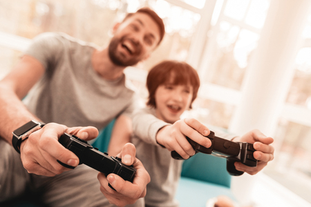 Father and Son Sitting and Playing on Console. Sitting Boy. Young Father. Playing on Console. Joystick in Hands. Young Bearded Man at Home. Sitting on Blue Sofa. Happy Family Concept.の写真素材