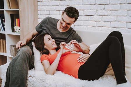 Couple is Relaxing on Couch. Couple is a Man and Pregnant Woman. Persons is Happy and Smiling. Woman is Showing a Heart Sign to Her Husband. Man is Touching a Woman's Belly. People Located at Home.の写真素材