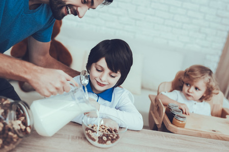 Man Spends Time with His Sons. Father of Boys is Engaged in Raising Children. Father is Feeding His Sons. Man is Pouring Milk to Cereal in Bowl. Boys is Sitting at Table. People Located on Kitchen.の写真素材