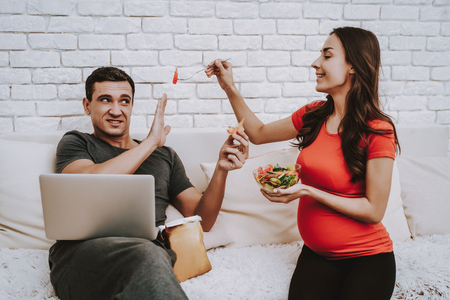 Couple is Sitting on Couch. Pregnant Woman is Giving a Salad to Husband. Salad in Bowl From Fresh Vegetables. Man Dont Want Eat a Salad. Man is Eating a Chips. Man is Holding a Laptop. People at Home.の写真素材