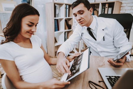 Pregnant Girl at the Gynecologist Doctor. Doctor is a Young Man. Doctor Showing the Ultrasound Results to Woman. People Sitting at Table. Persons is Smiling. People Located at Clinic.の写真素材