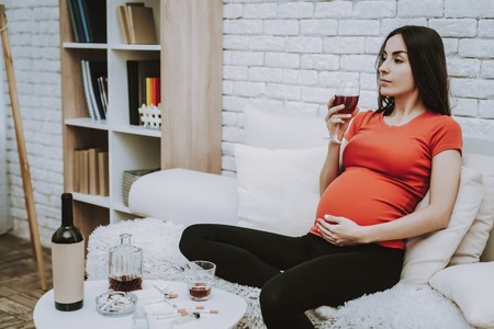 Woman Watching a TV. Woman is a Young Brunette Pregnant Girl. Girl is Sitting on Couch and Drinking a Wine. Woman is Touching Her Belly. Cigarettes and Alcohol on Table. Person Located at Home.の写真素材