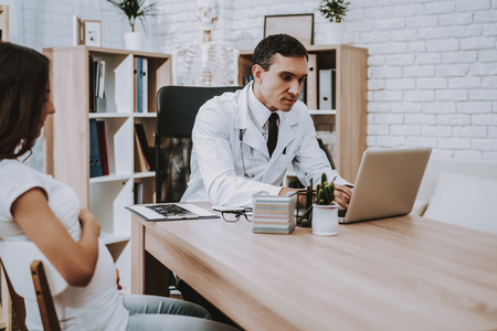 Pregnant Girl at the Gynecologist Doctor. Doctor is a Young Man. Girl is a Young Brunette Woman. Doctor is Typing Information about Woman in Laptop. People Sitting at Table. People Located at Clinic.の写真素材
