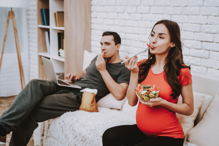 Couple is Sitting on Couch. Pregnant Woman is Eating a Salad with a Fork. Salad in Bowl From Fresh Vegetables. Man is Eating a Chips. Man is Using a Laptop. People Located at Home Interior.の写真素材