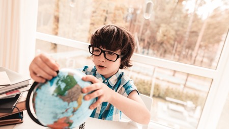 Sitting Little Boy in Glasses Studying Globus. Education at Home. White Table in Room. Sitting Boy....