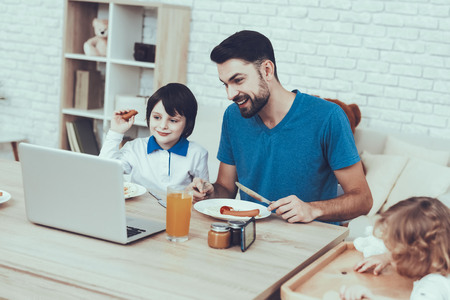 Man Spends Time with His Sons. Father is Engaged in Raising Children. Eldest Son and Father is Eating a Breakfast and Watching Video on Laptop. People is Sitting at Table. People Located at Home.の写真素材