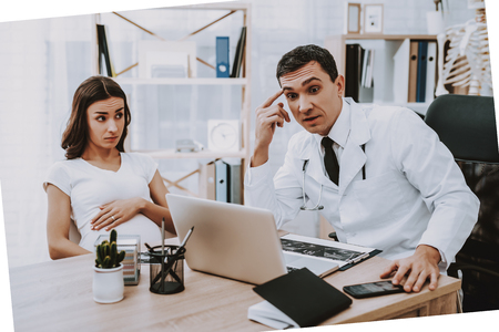 Pregnant Girl at the Gynecologist Doctor. Doctor is a Young Man. Woman is Touching Her Belly. Surprised Doctor is Looking on Laptop Screen. People Sitting at Table. People Located at Clinic.の写真素材