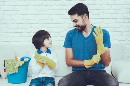 Man Spends Time with His Son. Father of Boy is Engaged in Raising Child. Father is Teaching a Son a Cleaning. Persons Weating a Gloves. Persons is Sitting on Couch. People is Located in Bedroom.の写真素材