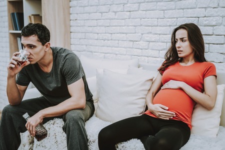 Couple Have a Conflict. Woman Feeling a Bad. Woman is a Young Pregnant Girl. Girl is Touching Her Belly. Man is Drinking a Whiskey. Upset People is Sitting on Couch. Persons Located at Home.の写真素材