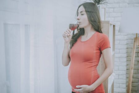 Woman is Looking out from Window. Woman is a Young Brunette Pregnant Girl. Girl is Drinking a Red Wine. Red Wine in Glass. Woman is Touching Her Belly. Person Located at Home Interior.の写真素材
