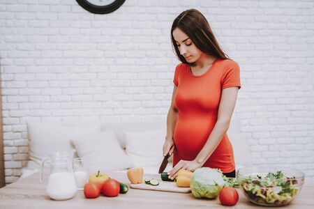 Woman is Cooking Salad from Fresh Vegetables. Woman is a Young Brunette Pregnant Girl. Girl is Cutting the Cucumber a Knife. Vegetables, Milk and Salad in Bowl on Table. Girl Located at Home Interior.の写真素材