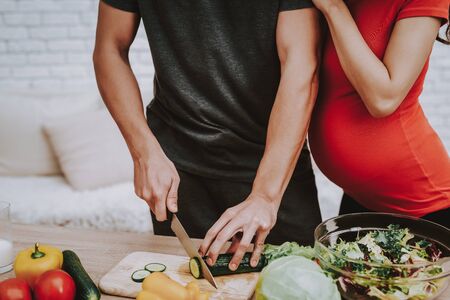 Man is Cooking Salad. Woman is Hugging Her Man. Woman is a Pregnant Girl. Man is Cutting the Cucumber a Knife. Vegetables, Milk and Salad in Bowl on Table. People Located at Home Interior.の写真素材