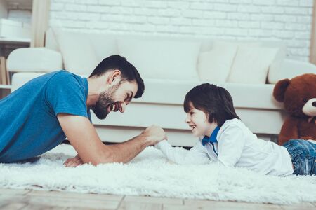 Man Spends Time with His Son. Father of Boy is Engaged in Raising Child. Father and Son is Playing in Arm wrestling. Persons is Lying on Carpet and Smiling. People is Located in Bedroom.の写真素材